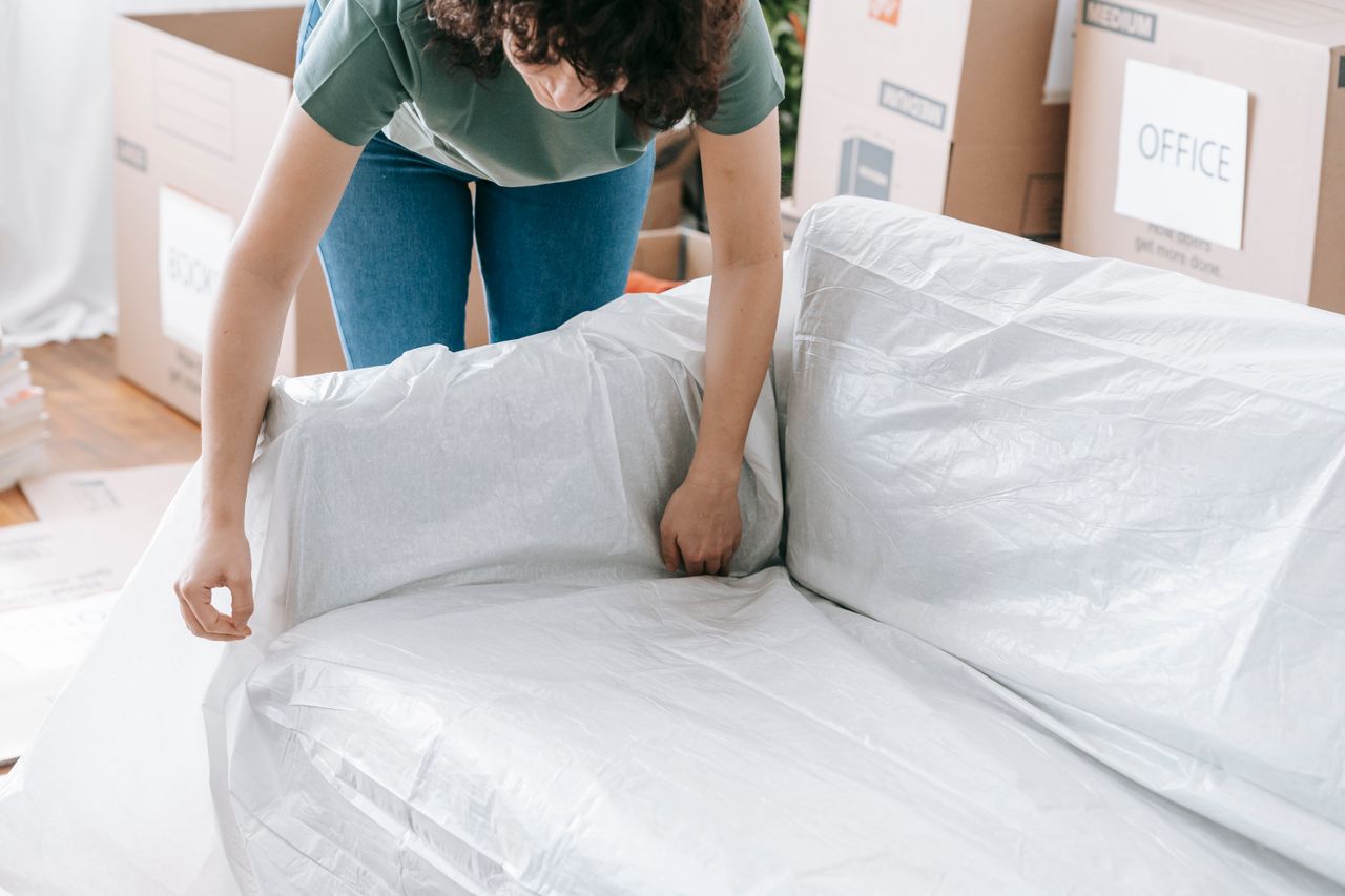 services-01 A woman carefully wraps a sofa in protective material while packing boxes in a bright room.